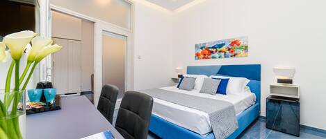 A modern bedroom with a blue bed, white walls, and a colorful painting above the bed.