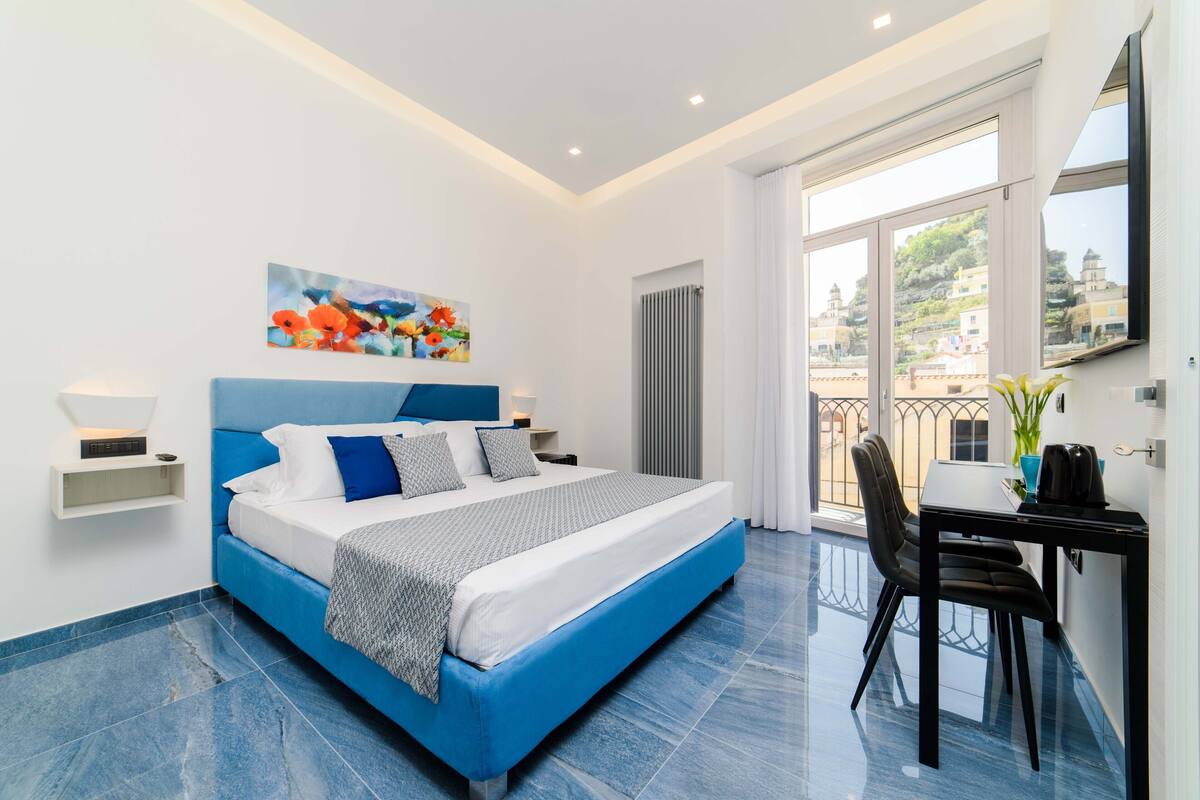 A modern bedroom with a large bed, a painting above the bed, a desk with a chair, and a balcony with a view of buildings.