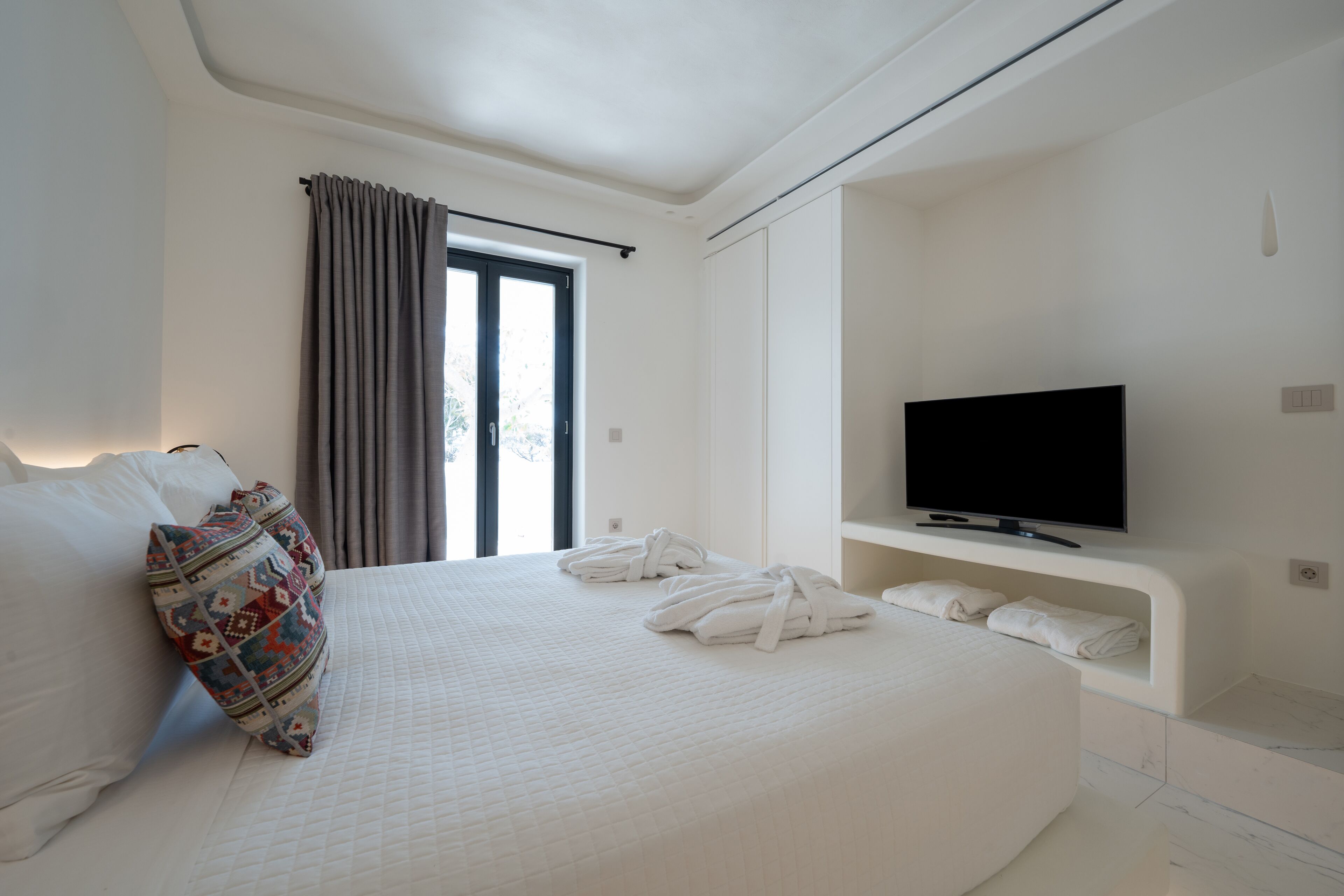 A hotel room with a large bed, a flat-screen TV mounted on the wall, and a window with curtains.