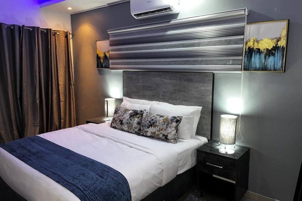 Una habitación de hotel moderna con una cama grande, una mesita de noche con lámpara, un equipo de aire acondicionado y un cuadro en la pared.