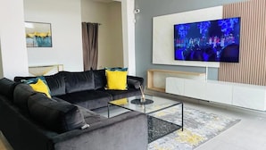 Ein modernes Wohnzimmer mit einem schwarzen Sofa, gelben Kissen, einem Glastisch und einem großen Flachbildfernseher an der Wand.