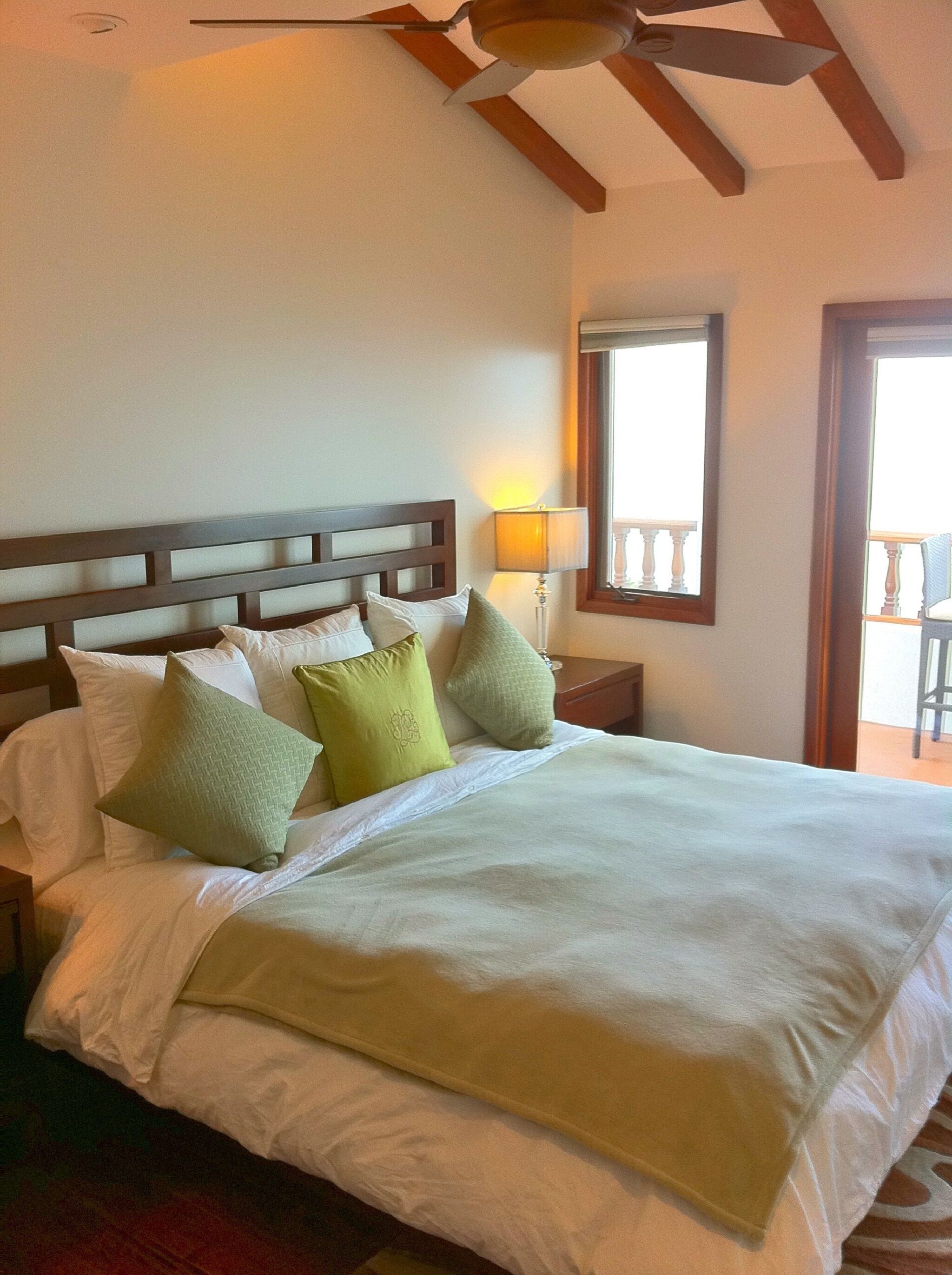 A neatly made bed with white linens and multiple pillows, a wooden headboard, a bedside lamp, and a view of a balcony through a window.