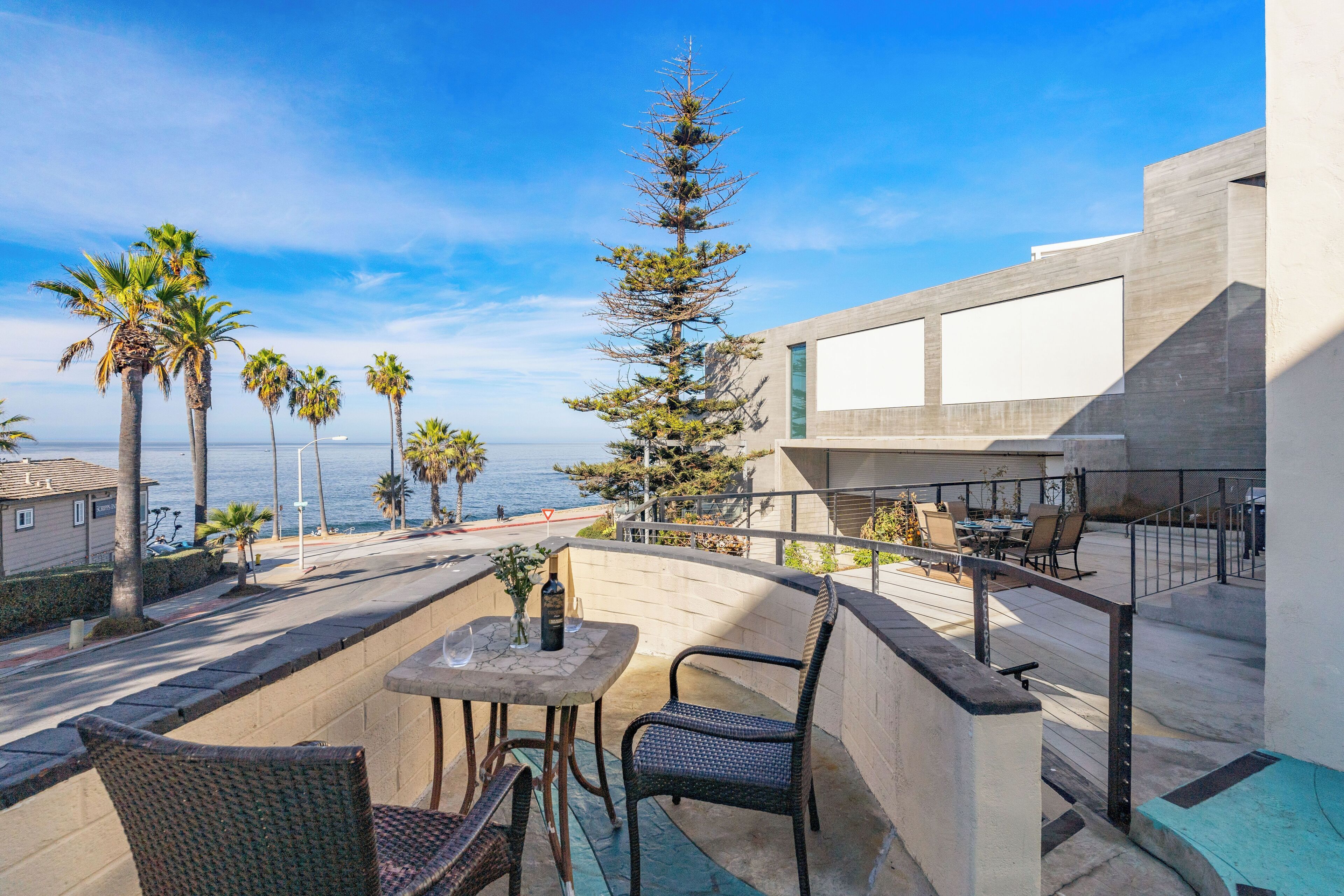 A balcony with a table and chairs, overlooking a coastal area with palm trees and a view of the sea.