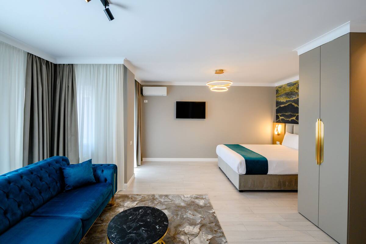 A modern hotel room with a blue sofa, a bed with a green throw, a flat-screen TV, and a marble-patterned rug.