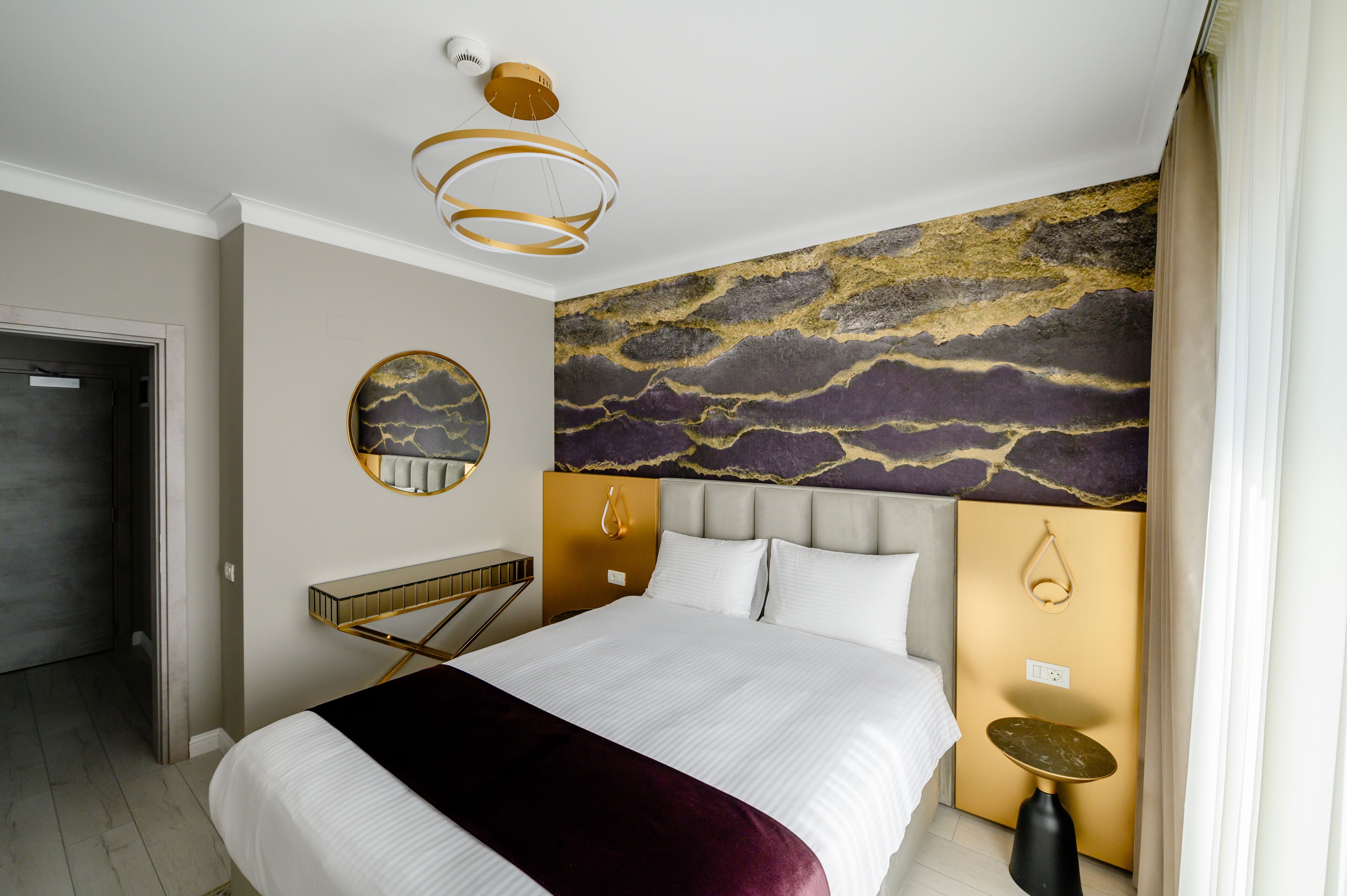 Una habitación de hotel moderna con una cama grande, un espejo redondo, un mural único en la pared y una lámpara de techo elegante.