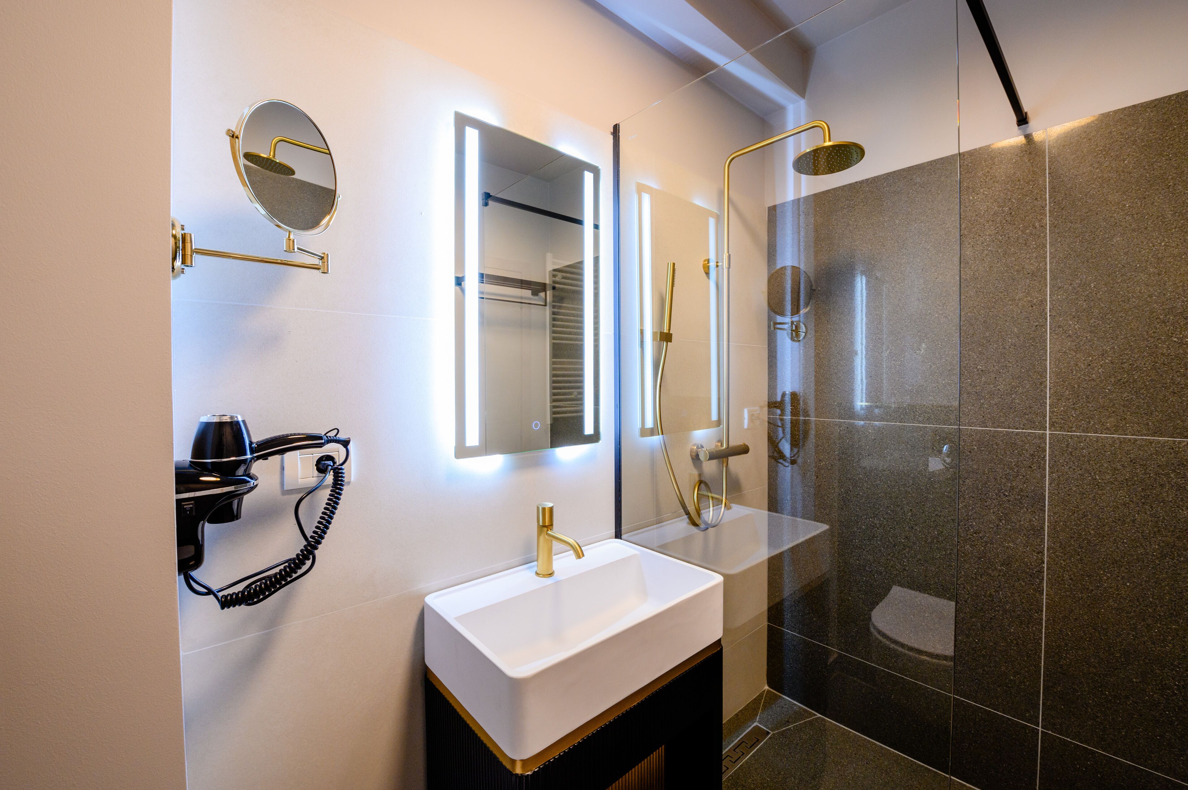 Een moderne badkamer met een witte wastafel, gouden details, een haardroger en een douche met glazen wanden.