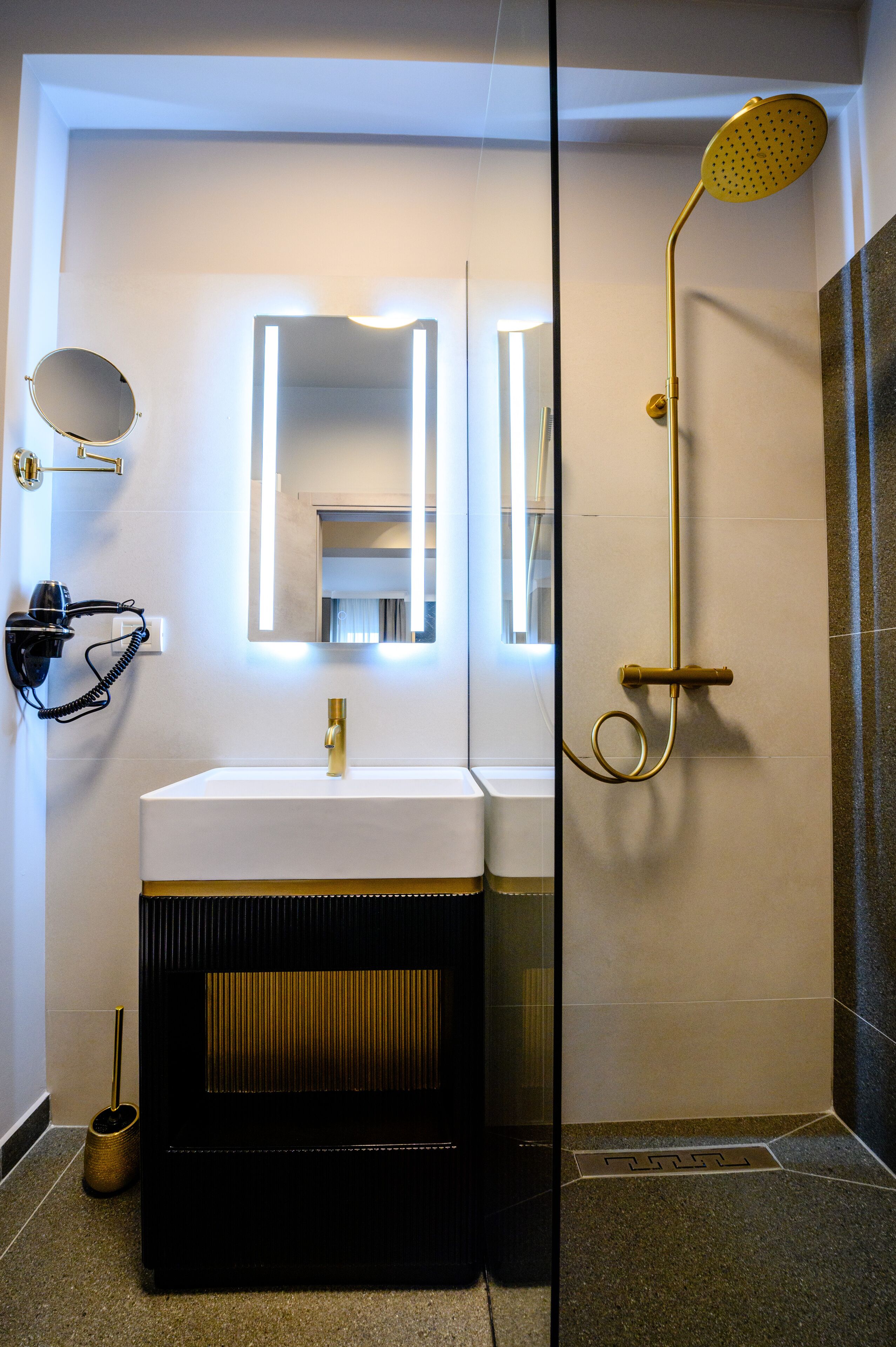 Une salle de bain moderne avec un lavabo blanc, des robinetteries dorées, une douchette à main et un miroir doté d’un éclairage intégré.