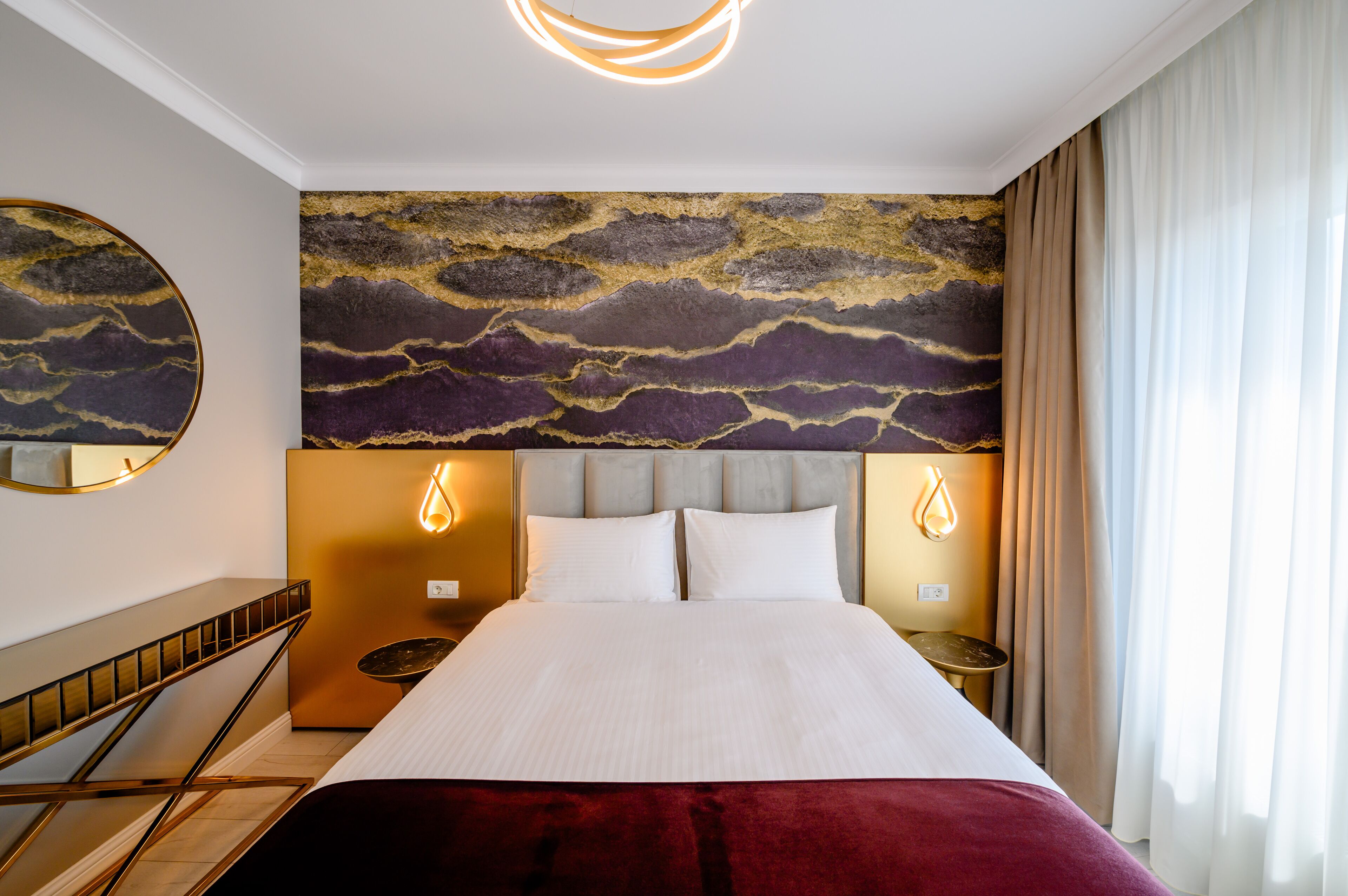 A hotel room with a large bed, a headboard with a textured wall design, two bedside tables with lamps, and a round mirror on the wall.