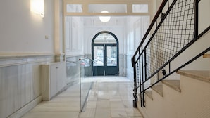 Um corredor com piso de mármore, guarda-corpo de vidro, escada de metal preta e uma porta com painel de vidro.