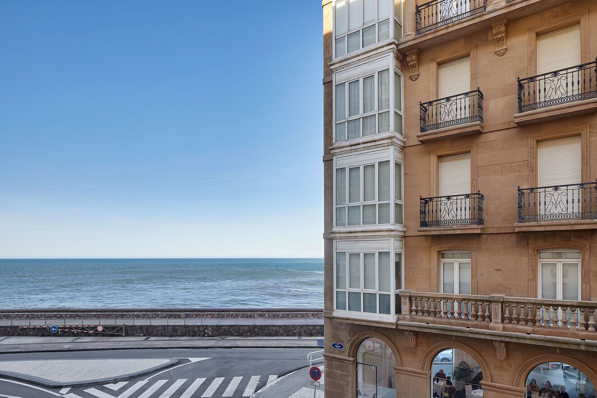 Un bâtiment situé sur la côte, doté de nombreux balcons, offrant une vue dégagée sur la mer et bordé d’une promenade piétonne au bord de l’eau.