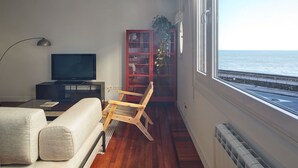 Uma sala de estar com sofá, televisão, cadeira de madeira, armário vermelho e vista para o mar pela janela.