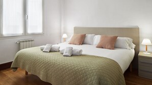 A neatly made bed with a quilted green bedspread, two bedside lamps, and a radiator on the wall.