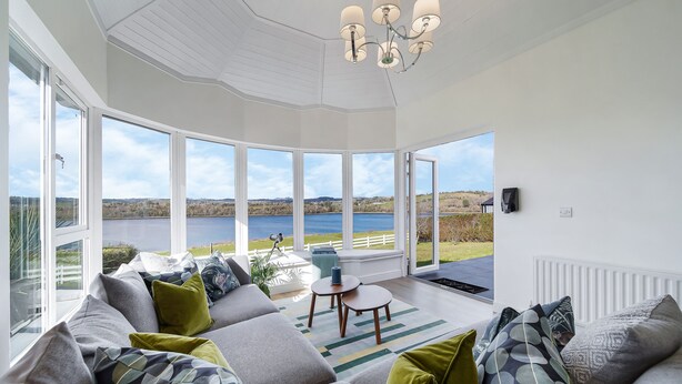 A modern living room with a large window overlooking a lake, furnished with a sofa and a coffee table. - Fresh & Stylish | Ocean View | Donegal Town & Bay (Tullycullion)