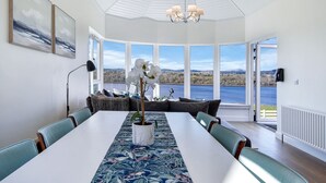 A dining area with a long table, chairs, a chandelier, and a view of a lake. - Fresh & Stylish | Ocean View | Donegal Town & Bay (Tullycullion)