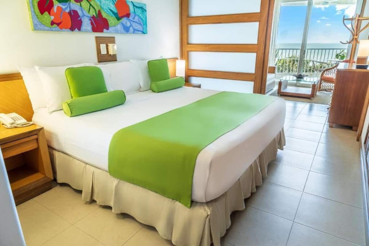 A hotel room with a large bed, green pillows, a telephone on the nightstand, and a balcony with a view.