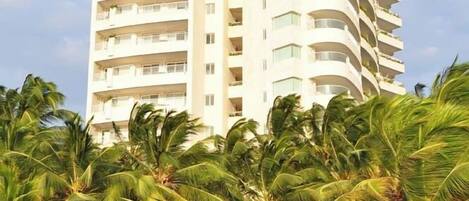 Un hotel de gran altura con varios balcones, rodeado de palmeras y una estructura con techo de paja.