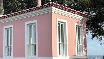 A pink building with white trim and multiple windows, surrounded by trees and a view of the sea.