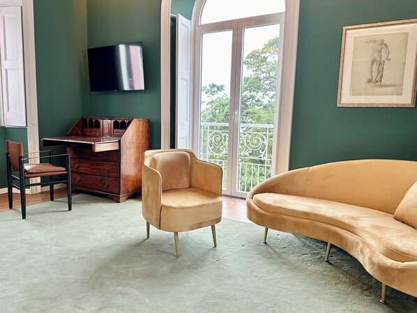 Una habitación con pared verde, un escritorio con silla, un sofá, un televisor y un balcón con vistas a árboles.