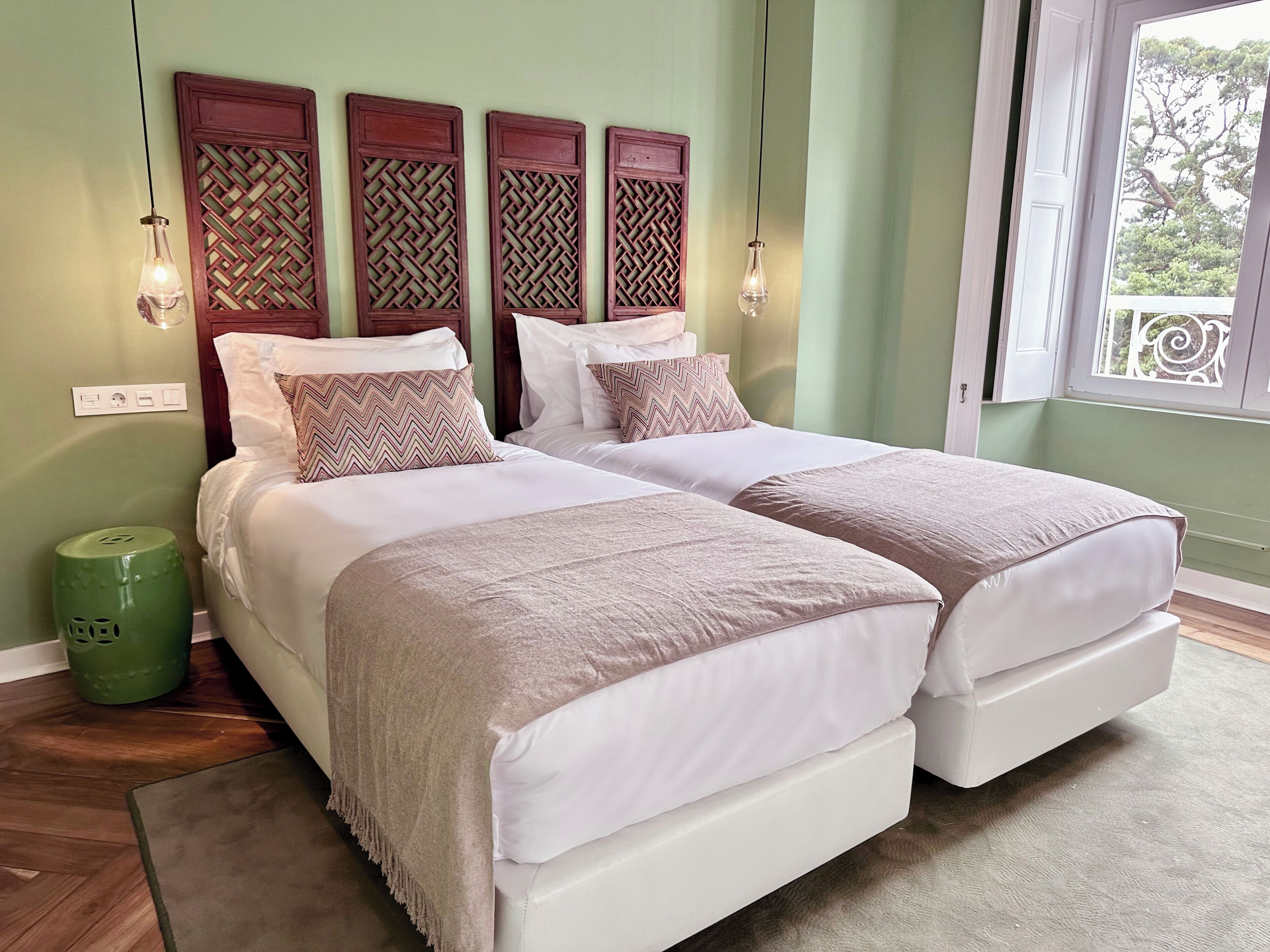 Una cama doble con dos almohadas, un cubo de basura verde y una ventana con vistas a árboles.