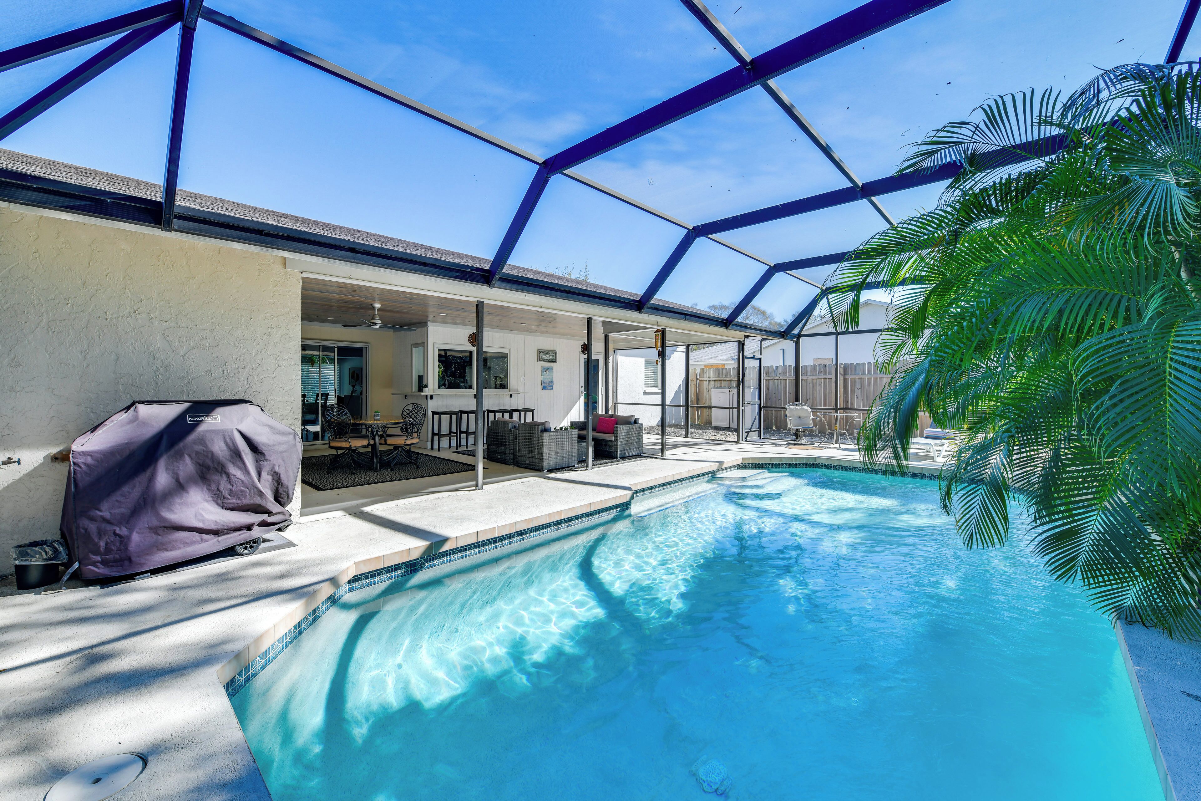 A covered outdoor swimming pool with a patio area, a covered grill, and a view of a house in the background.