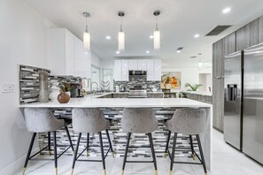 Une cuisine moderne dotée d’un îlot central, de tabourets de bar, d’appareils électroménagers en acier inoxydable et de luminaires suspendus.