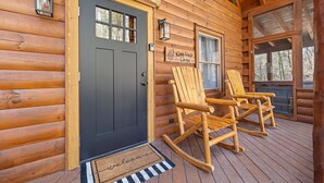 Une cabane en rondins avec une porte d’entrée sombre, deux fauteuils à bascule et un paillasson.