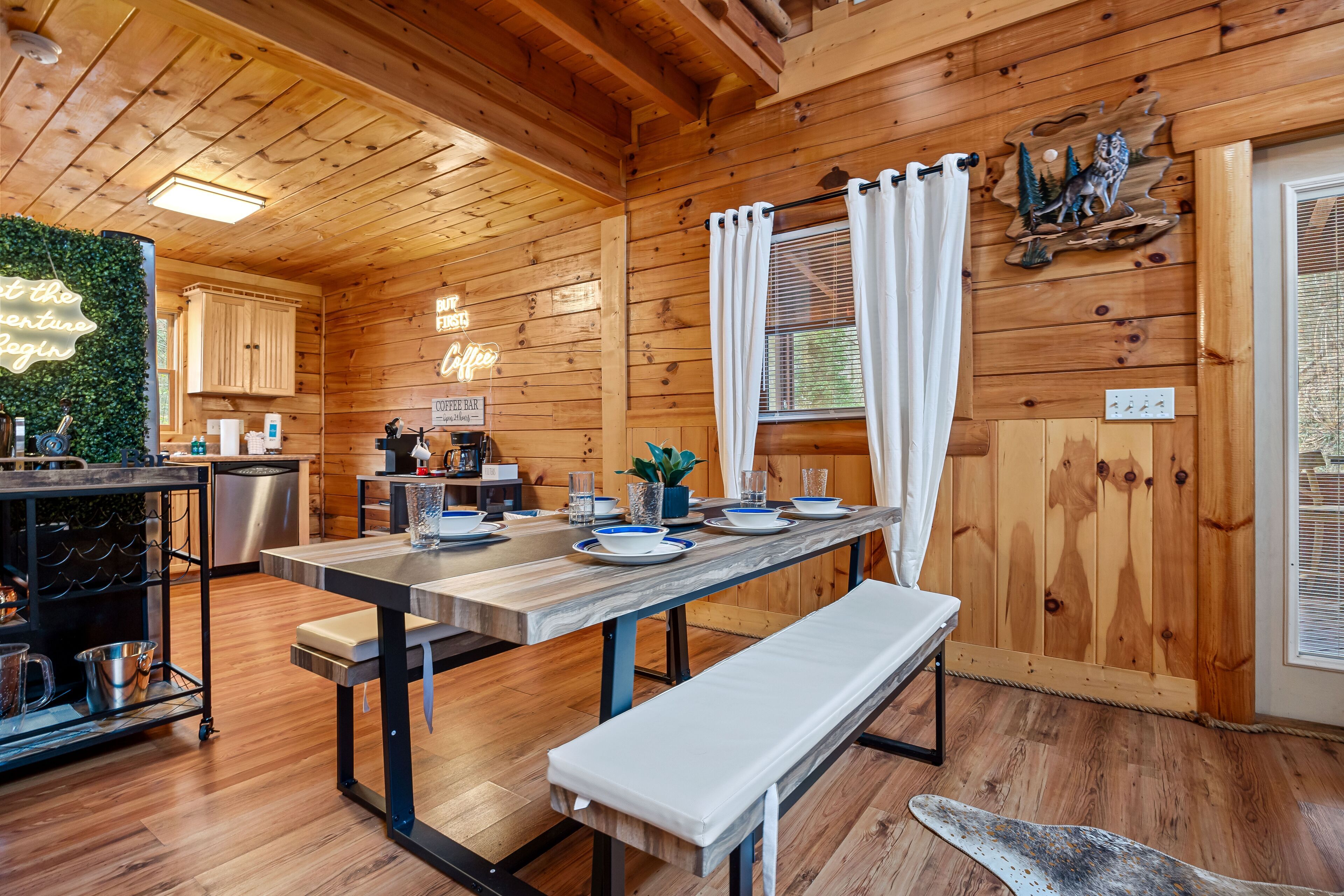 Interior de una cabaña de madera con una mesa de comedor preparada para cuatro personas, un banco y una zona de cocina con una barra para café.