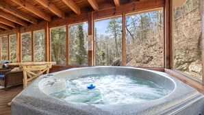 Un bain à remous en pierre, avec un flotteur bleu, entouré de meubles en bois et doté de grandes fenêtres donnant sur une zone boisée.