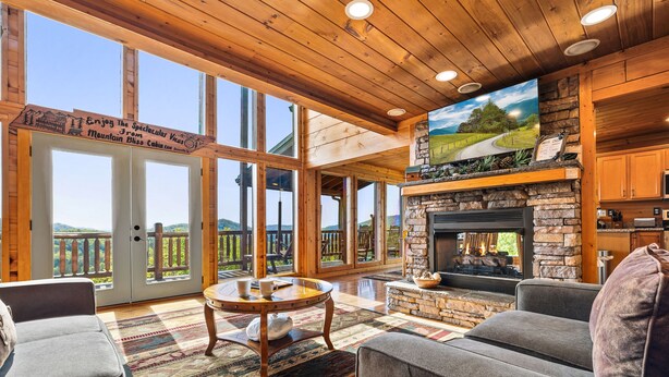 A spacious living room with a stone fireplace, large windows, and a view of the mountains. - The Joy Of The Mountain Tops (SEVIERVILLE)