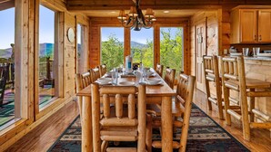 A wooden dining room with a large table and chairs, large windows with a view of greenery, and a chandelier. - The Joy Of The Mountain Tops (SEVIERVILLE)