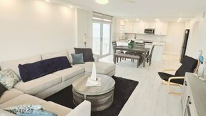 Een moderne woonkamer met een bank, salontafel en televisie. Achtergrond toont een keuken met een eethoek.