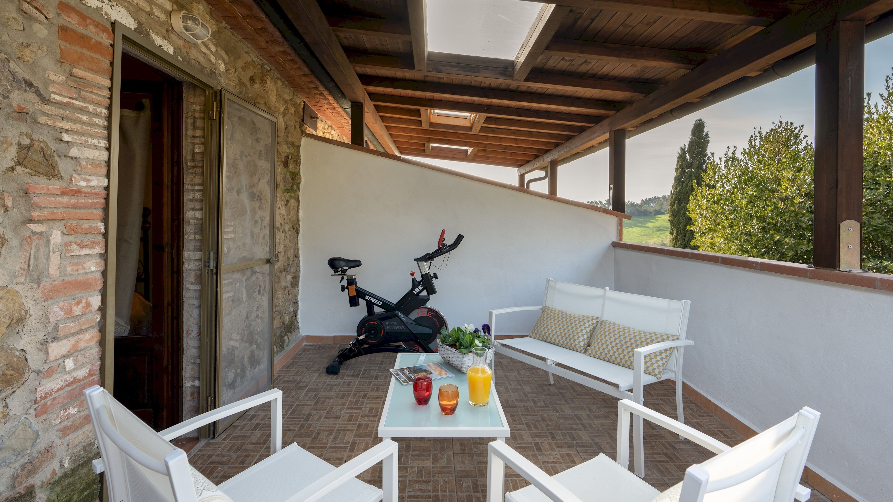 A covered patio with a brick wall, a white bench, a table with a basket, a bicycle, and a view of greenery.
