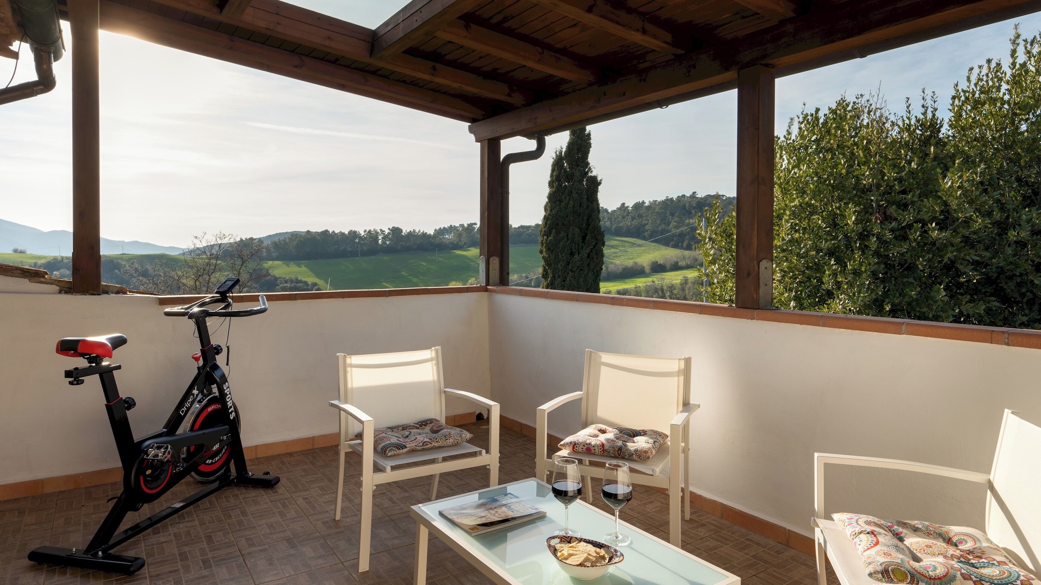A balcony with a stationary exercise bike, two white chairs, a glass table with a bowl and wine glasses, and a scenic view of green fields and trees.