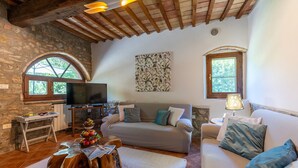 A cozy living room with a stone wall, wooden ceiling beams, a flat-screen TV, a sofa, a coffee table, and a lamp. - Il Pratone 12, Emma Villas (Pomarance)