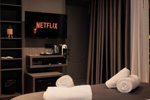 Una habitación de hotel con una televisión de pantalla plana mostrando el logotipo de Netflix, un minibar, una cafetera y toallas ordenadamente dobladas en la cama.