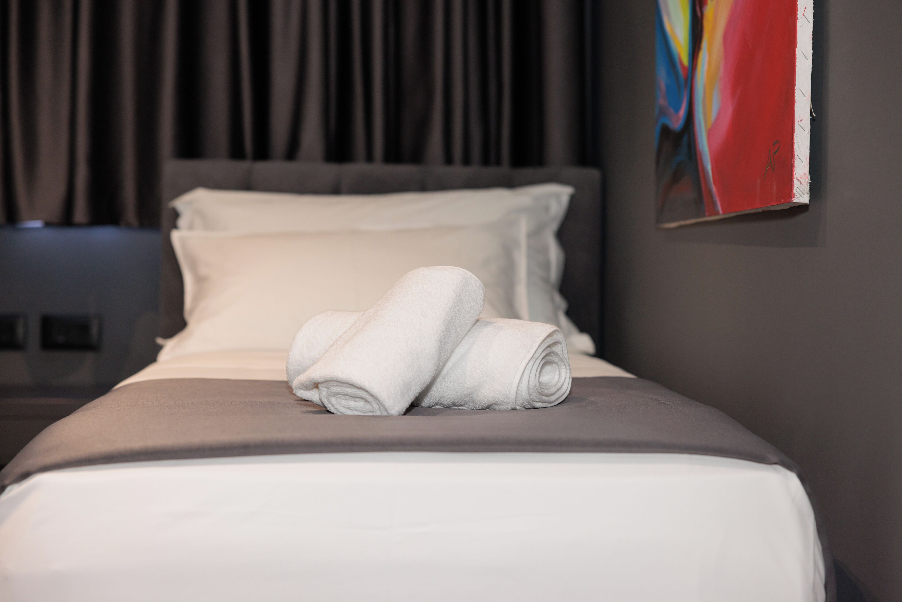 A neatly made bed with white linens and pillows, a gray headboard, and a colorful abstract painting on the wall.