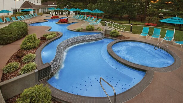Piscina de varios niveles con sillas de descanso y sombrillas, rodeada de zonas ajardinadas y un edificio al fondo.