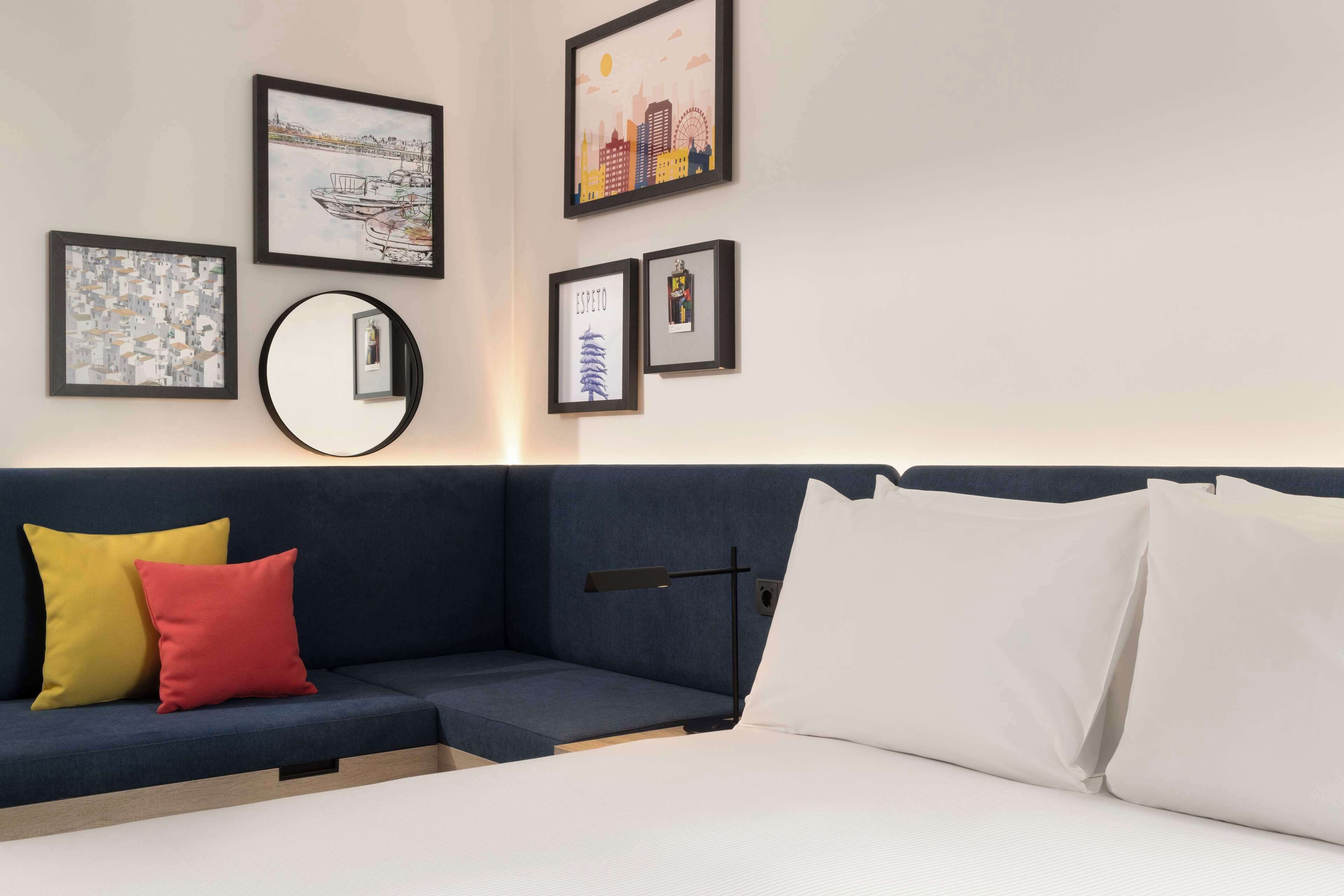 A modern hotel room with a dark blue sofa, a round mirror, and framed artwork on the wall.