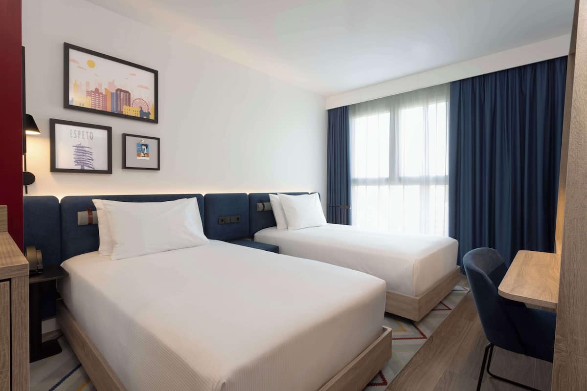 Quarto de hotel com duas camas, uma escrivaninha e uma cadeira. Quadros emoldurados decoram a parede.