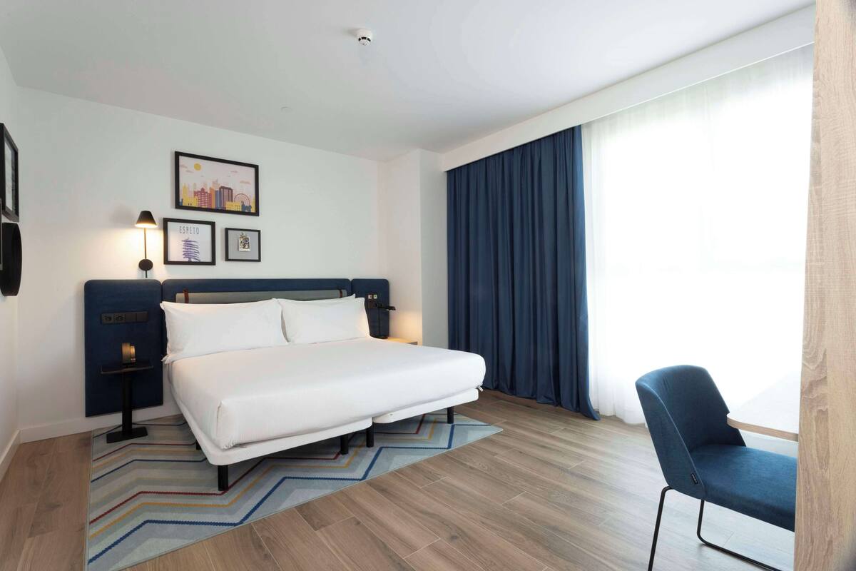 Quarto de hotel moderno com uma cama grande, uma mesa de cabeceira, uma cadeira e uma janela ampla com cortinas.