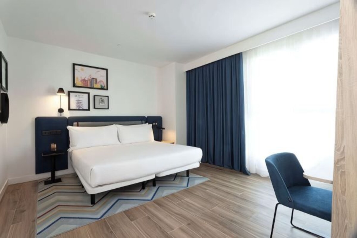 Habitación de hotel con cama, escritorio, silla, mesita de noche, lámpara y ventana con cortinas.