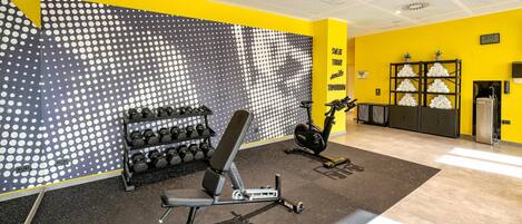 Ein Fitnessraum mit Wand voller kreisrunder Muster, einer Bank, Kurzhanteln, einem Ergometer und einer Uhr.
