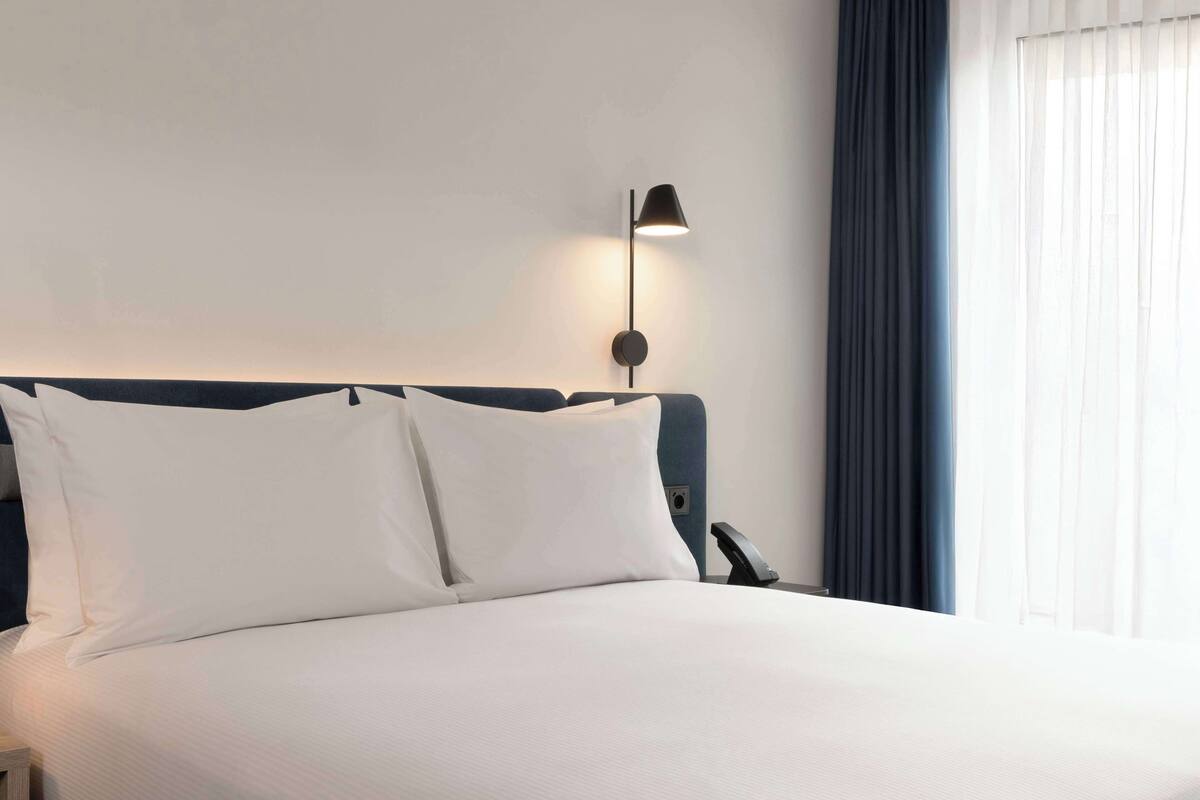 Cama bem arrumada com lençóis brancos e cabeceira, mesa de cabeceira com abajur e janela com cortinas.
