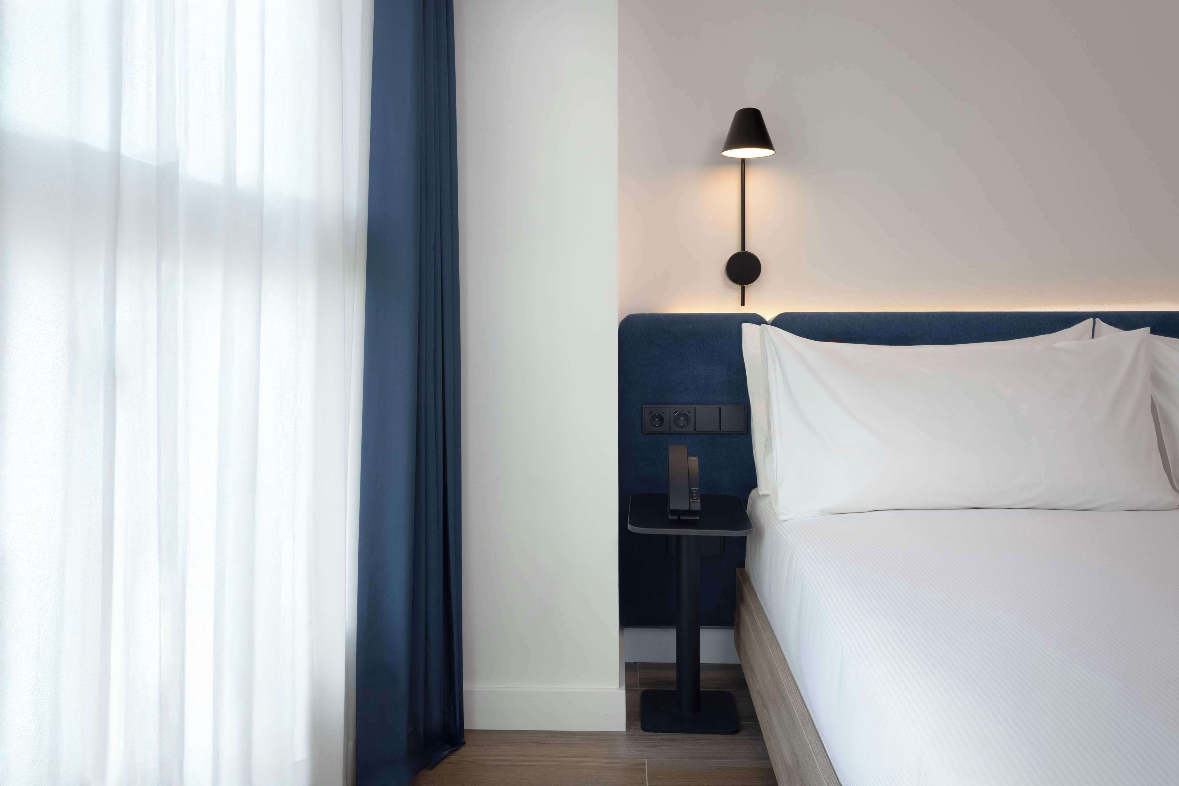 A hotel room with a bed, a nightstand with a lamp, and a window with curtains.