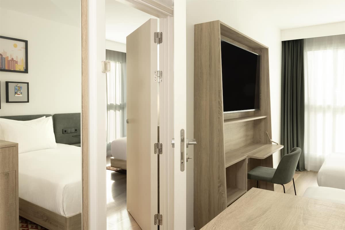Quarto de hotel com uma cama, uma escrivaninha com cadeira, uma televisão e um guarda-roupa.
