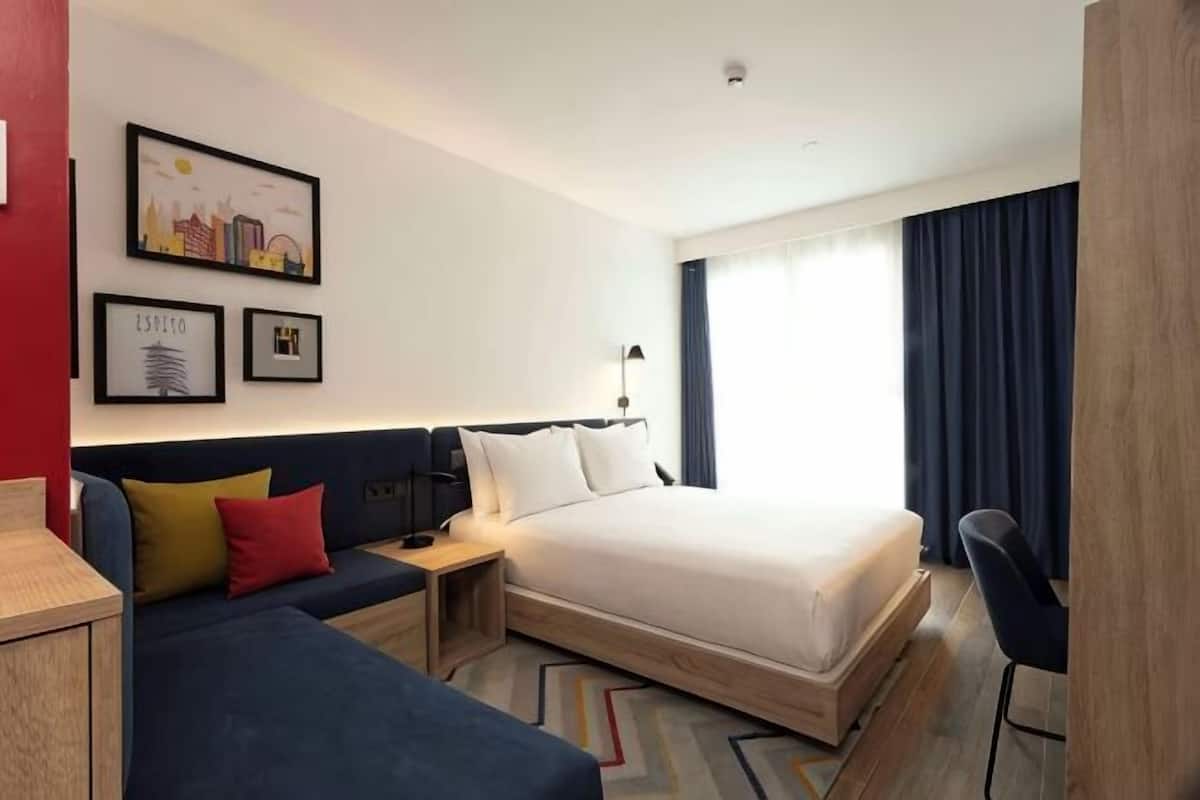 Quarto de hotel com cama, sofá, escrivaninha e cadeira. Quadros emoldurados decoram a parede.