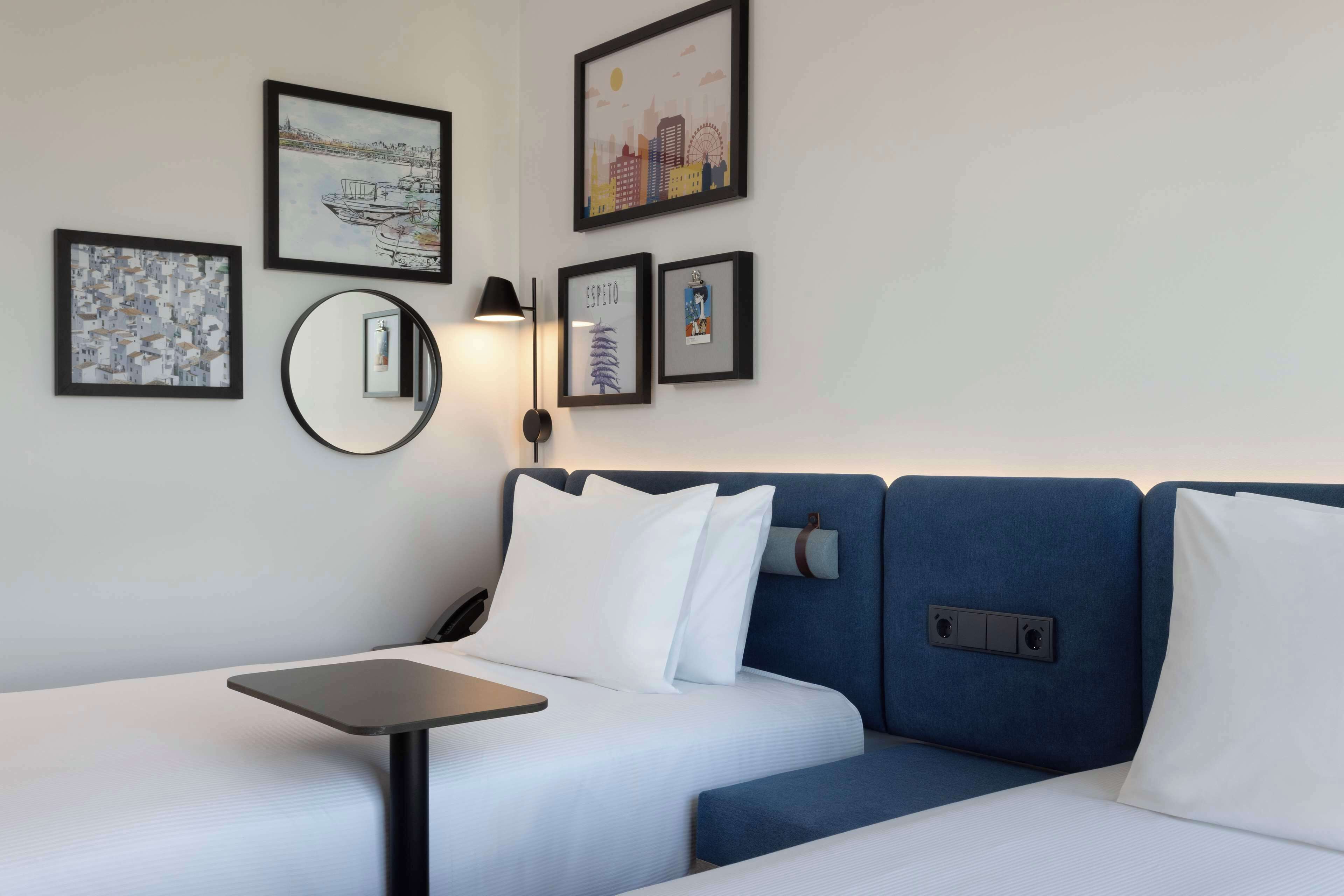 Habitación de hotel con cama, una mesita y un espejo montado en la pared. Hay cuadros enmarcados en la pared.