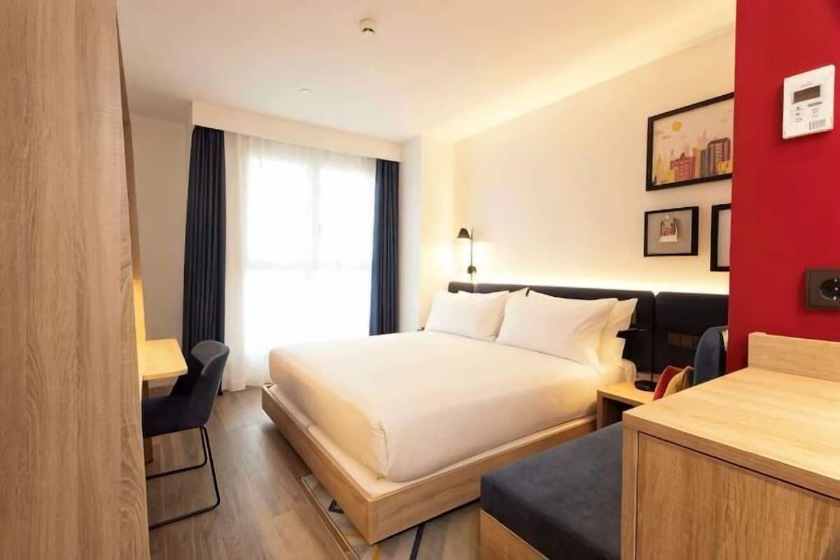 Quarto de hotel com cama, escrivaninha, cadeira, mesa de cabeceira e parede de destaque vermelha.