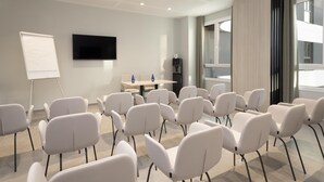 Une salle de conférence avec des chaises blanches disposées en rangées, un tableau blanc, un téléviseur à écran plat, une table avec deux bouteilles d’eau et une fenêtre avec des rideaux.