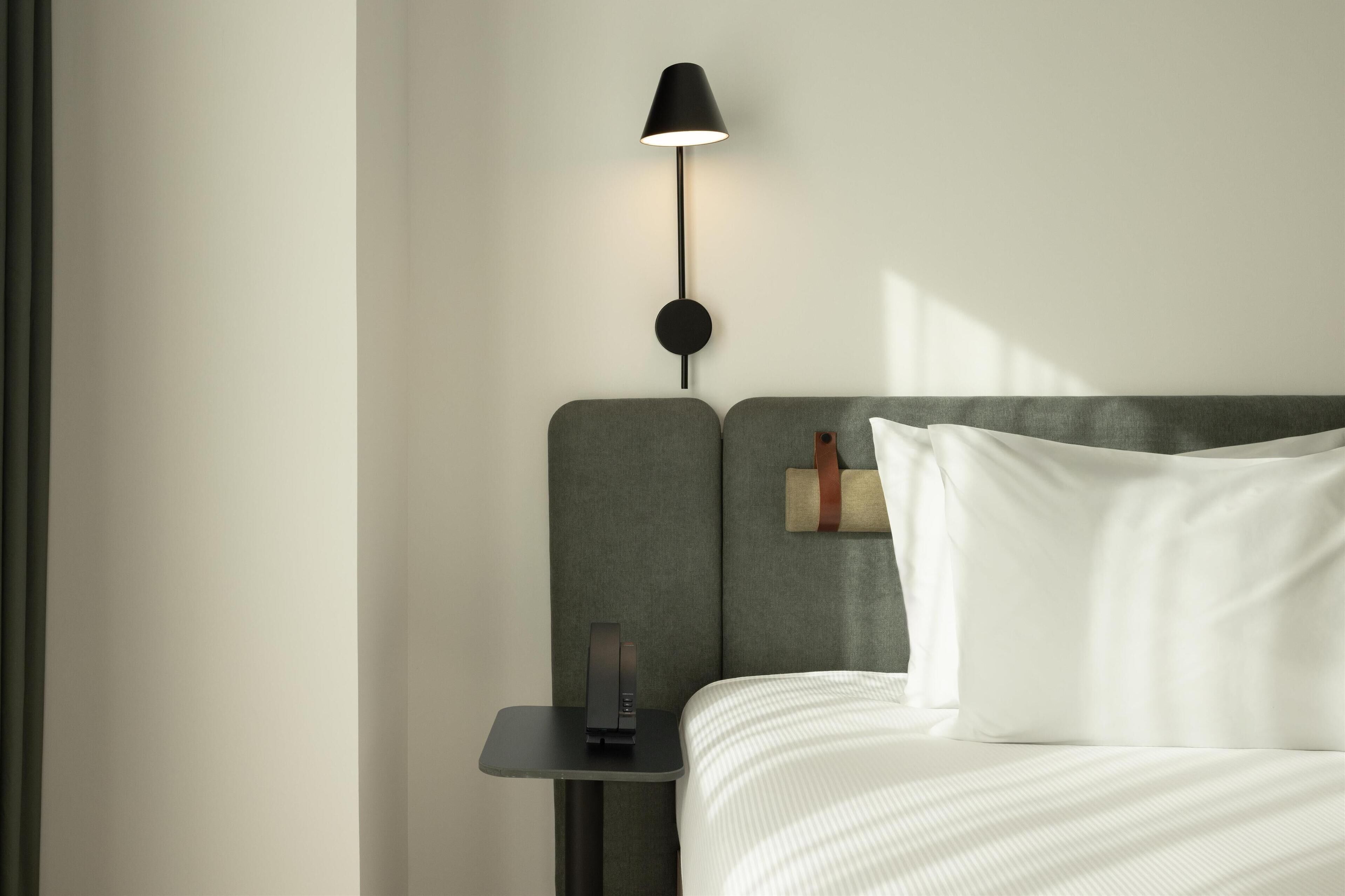 Una cama bien hecha con ropa blanca, una mesita de noche pequeña con una lámpara y un reloj de pared.